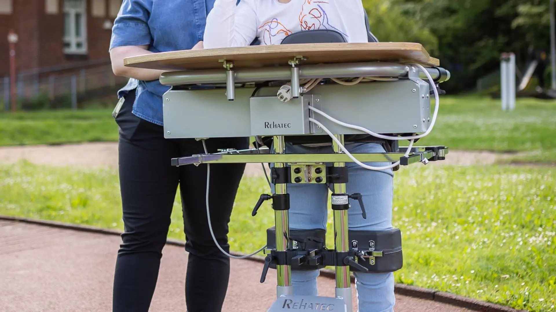 Eine Physiotherapeutin spielt mit einem Kind in einem Stehständer draußen auf dem Schulgelände. Beide lachen und haben Spaß am Spiel.