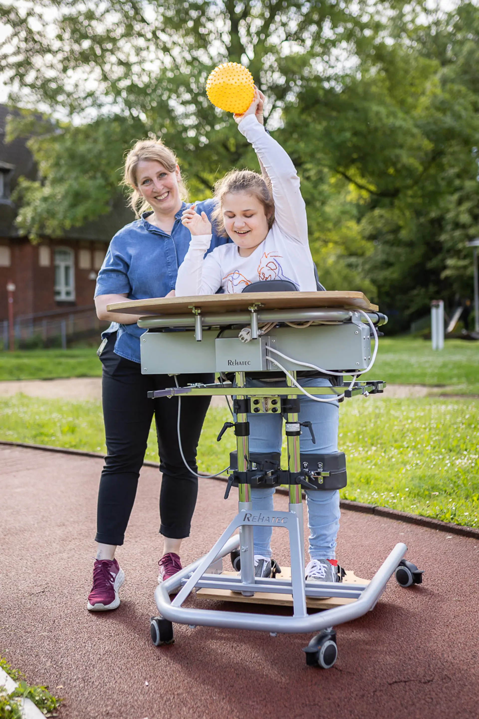 Eine Therapeutin und ein Kind im Stehstuhl spielen mit einem Ball und lachen draußen auf dem Schulgelände.