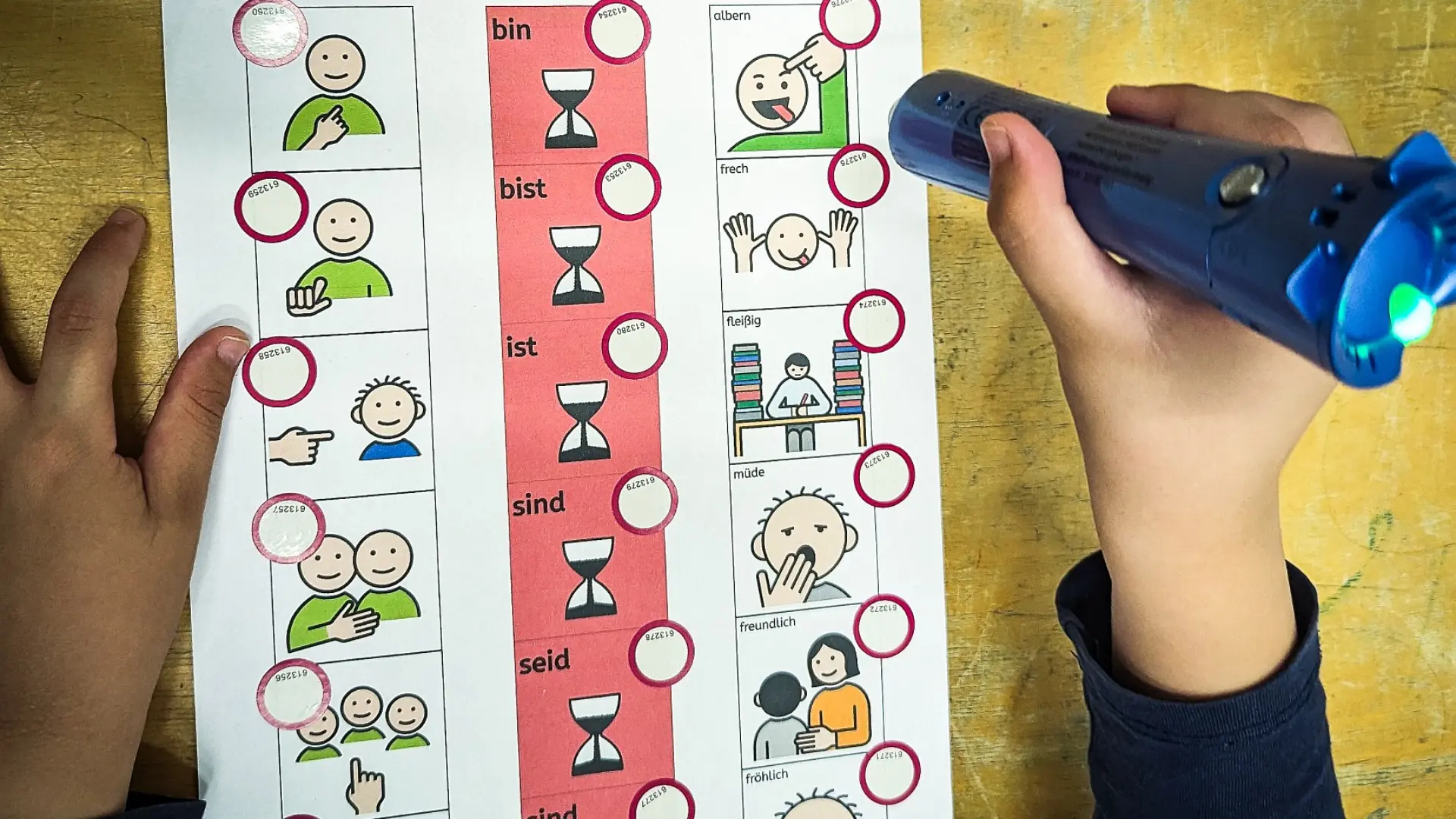 Die Hände eines Kindes: Mit der einen Hand hält es ​​​​​einen Audiostift, mit der anderen ein Satzleistenarbeitsblatt, worauf verschiedene Metacom-Symbole zu sehen sind. Mit beiden Hilfsmitteln kann das Kind verschiedene Satzstrukturen ausprobieren und erlernen. 