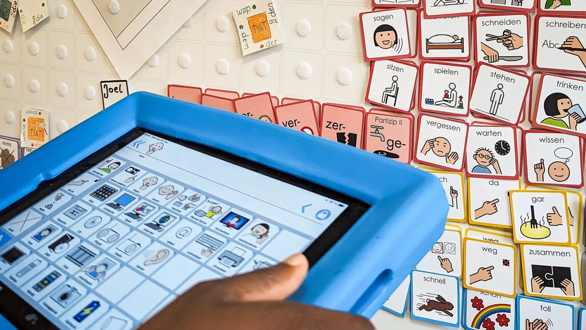 Die Hände eines Kindes, das ein Tablet hält, worauf verschiedene Metacom-Symbole zu sehen sind. Das Tablet ist auch zu einer Magnetwand gerichtet, woran viele einzelne Metacom-Symboltäfelchen zur Auswahl befestigt stehen. 