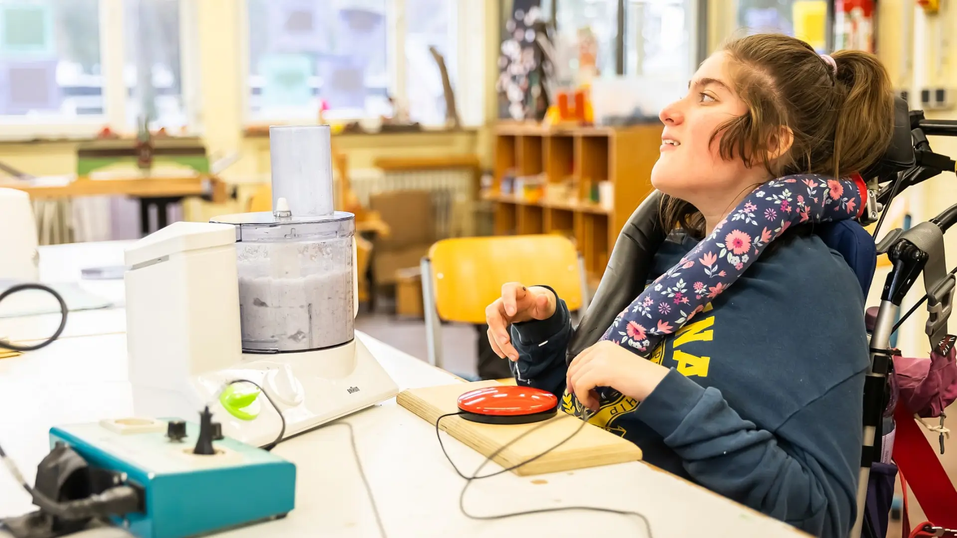 Eine Schülerin mit komplexer Beeinträchtigung ist im Rollstuhl und sitzt vor einem Arbeitstisch im Klassenraum. Vor ihr ist ein Mixer, den sie per Drücken eines großen Plastikknopfs bedienen kann.