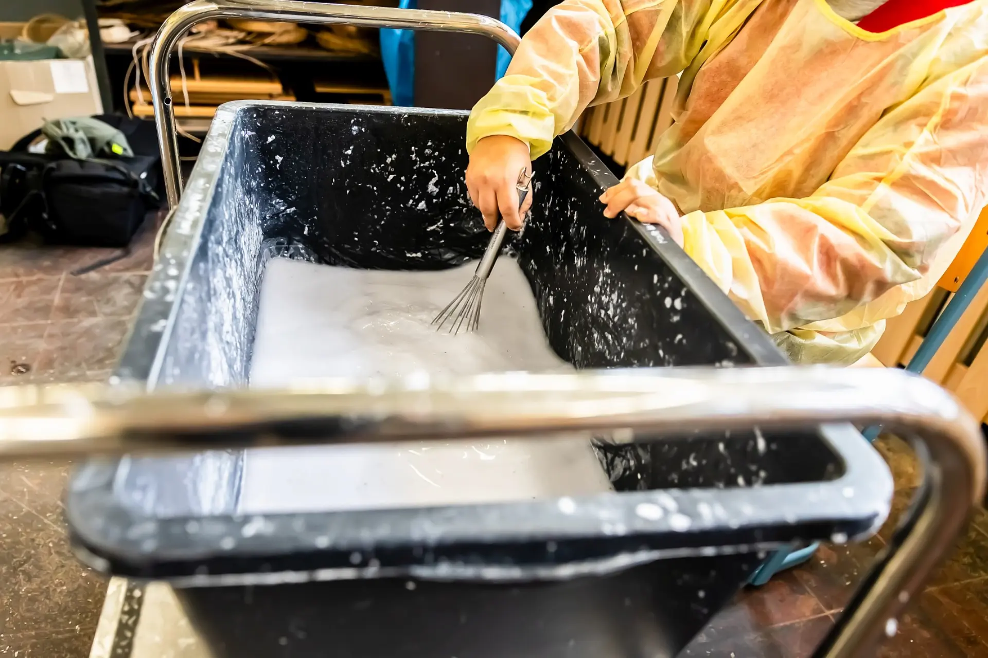 Ein etwa einen Meter großer Plastikbecken, der zur Hälfte mit einer feinen dickflüssigen Masse aus Papier und Wasser voll ist. Ein Kind mit Schürze steht am Rande des Beckens und rührt die Masse mit einem Handrührstab um.