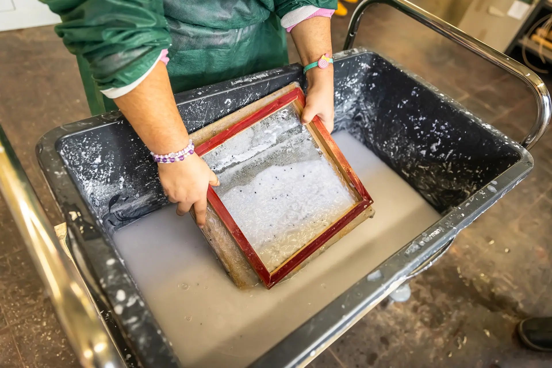 Ein Schulkind steht vor einem großen Plastikbecken mit der Papier-Wasser-Flüssigkeit, nimmt mithilfe eines Siebs ein Teil der aufgeweichten Papiermasse heraus und tropft es ab. 