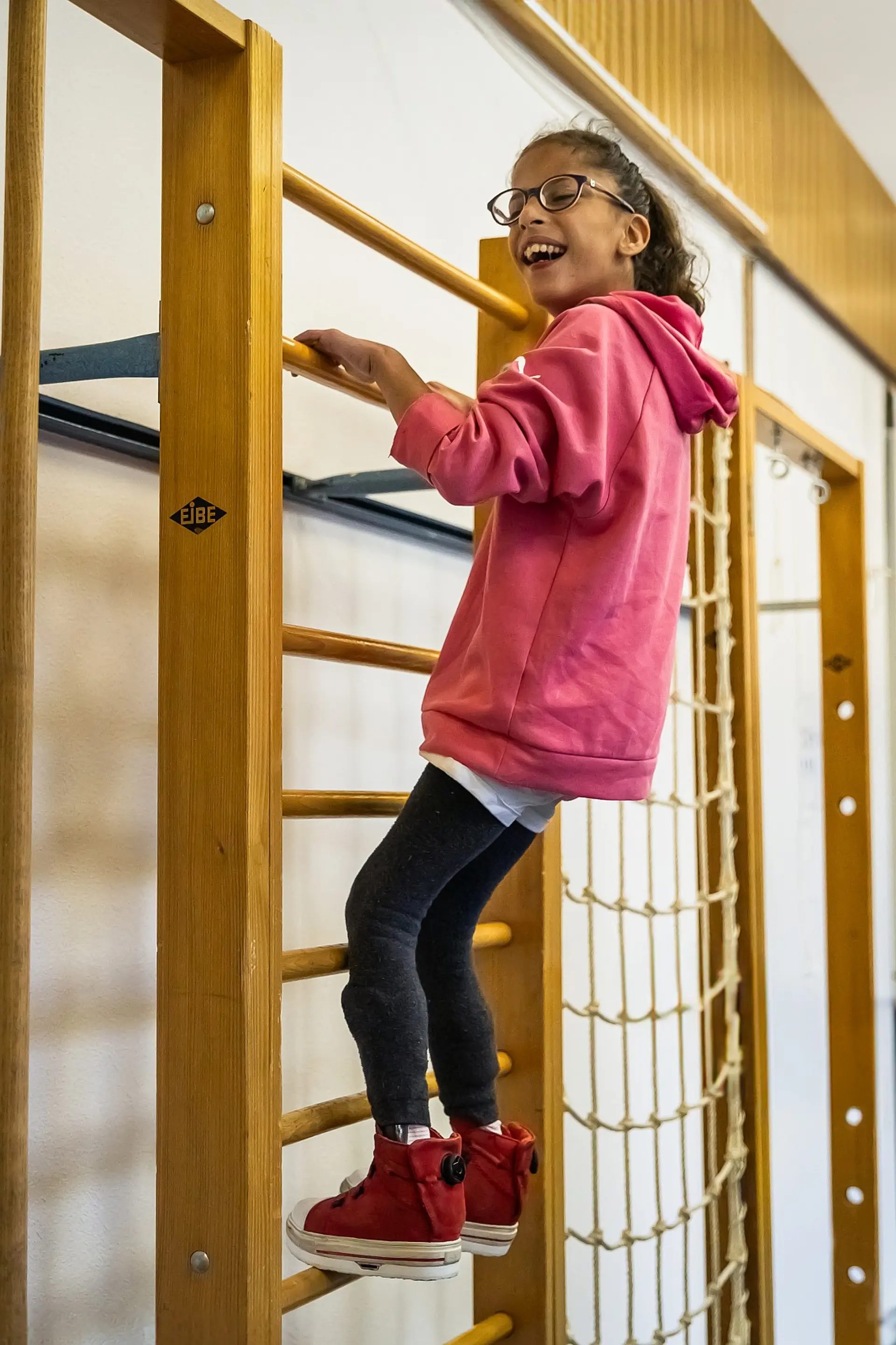 Eine Schülerin mit komplexer Beeinträchtigung klettert an einer Sprossenwand in der Sporthalle. Sie ist ganz oben gelangen und lacht.