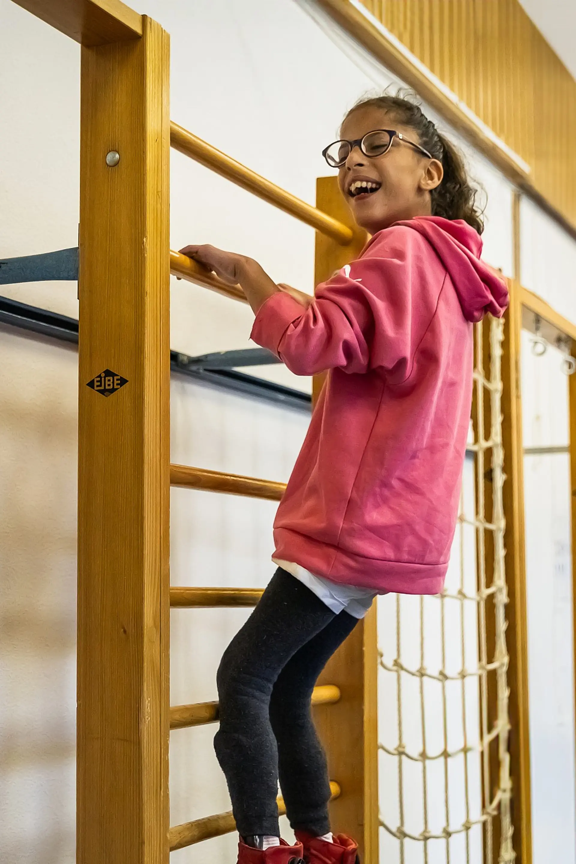 Eine Schülerin mit komplexer Beeinträchtigung klettert an einer Sprossenwand in der Sporthalle. Sie ist ganz oben gelangen und lacht.