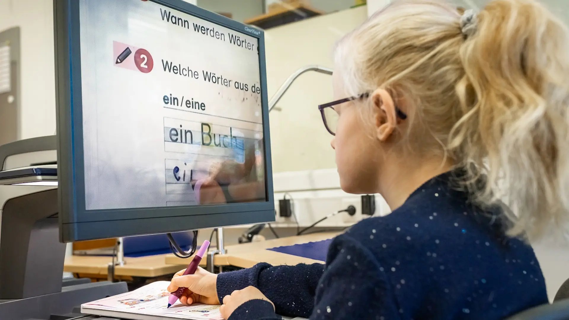Eine Schülerin sitzt an ihrem Arbeitsplatz im Klassenzimmer und macht Schreibübungen. Sie schreibt in ein spezielles Heft und sieht das Geschriebene stark vergrößert an einem Computerbildschirm vor ihr.