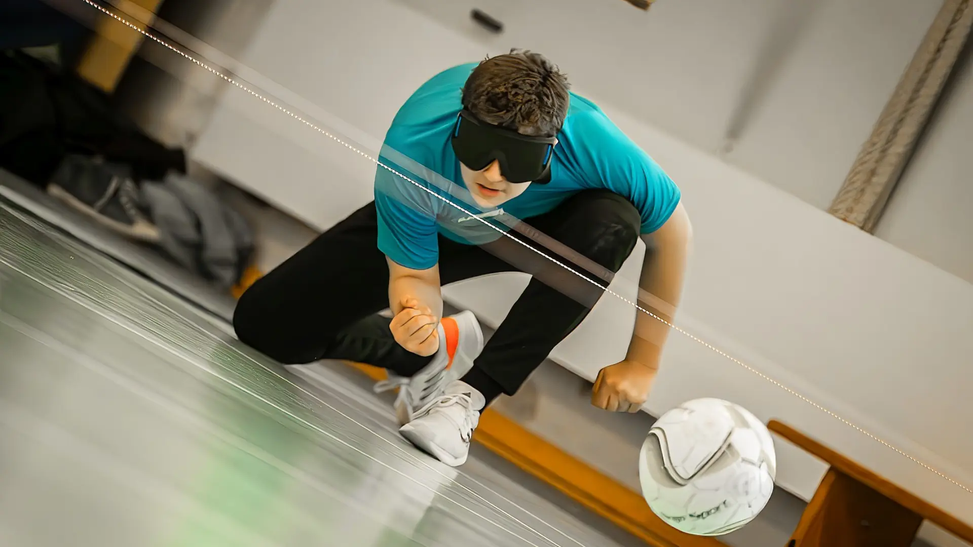 Schüler spielt Torball in einer Sporthalle. Er trägt eine große schwarze Brille, kniet auf dem Boden und hat gerade den Ball nach vorne geworfen. Der nach vorne fliegende Ball ist auch zu sehen. 