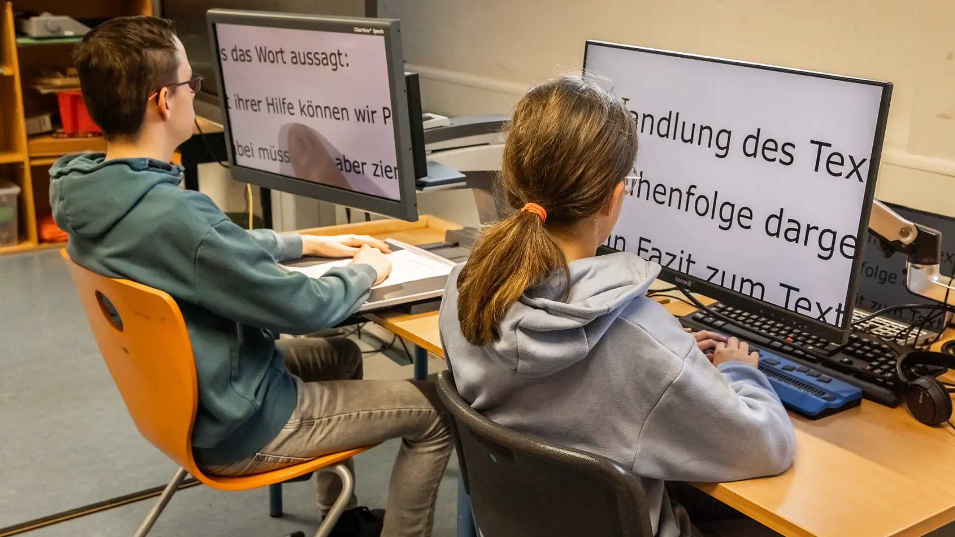 Zwei Schüler*innen sitzen am Schreibtisch in einem Schulraum. Vor jedem Jugendlichen sind jeweils ein Bildschirmlesegerät und eine Braillezeile, mit denen sie Texte lesen.