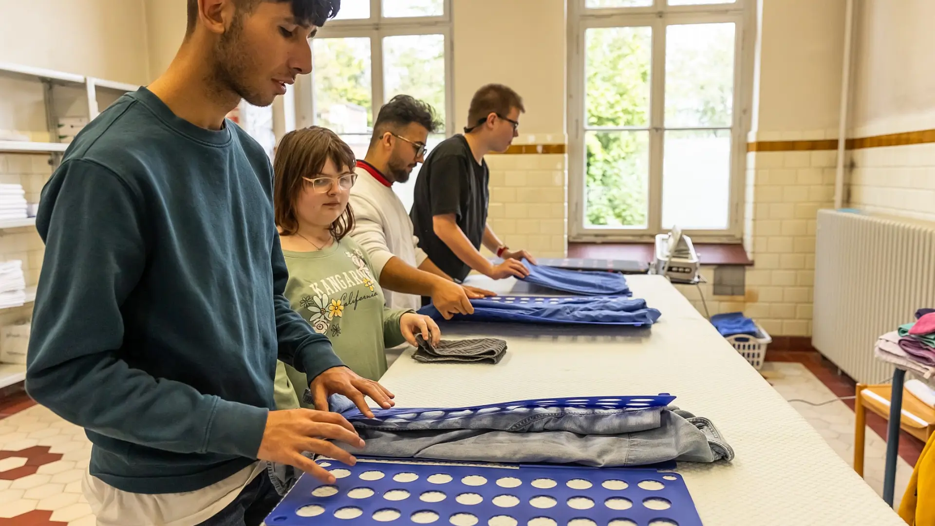 Vier Schüler*innen stehen vor einem langen Tisch und falten Wäsche mithilfe von Wäschefaltbrettern.