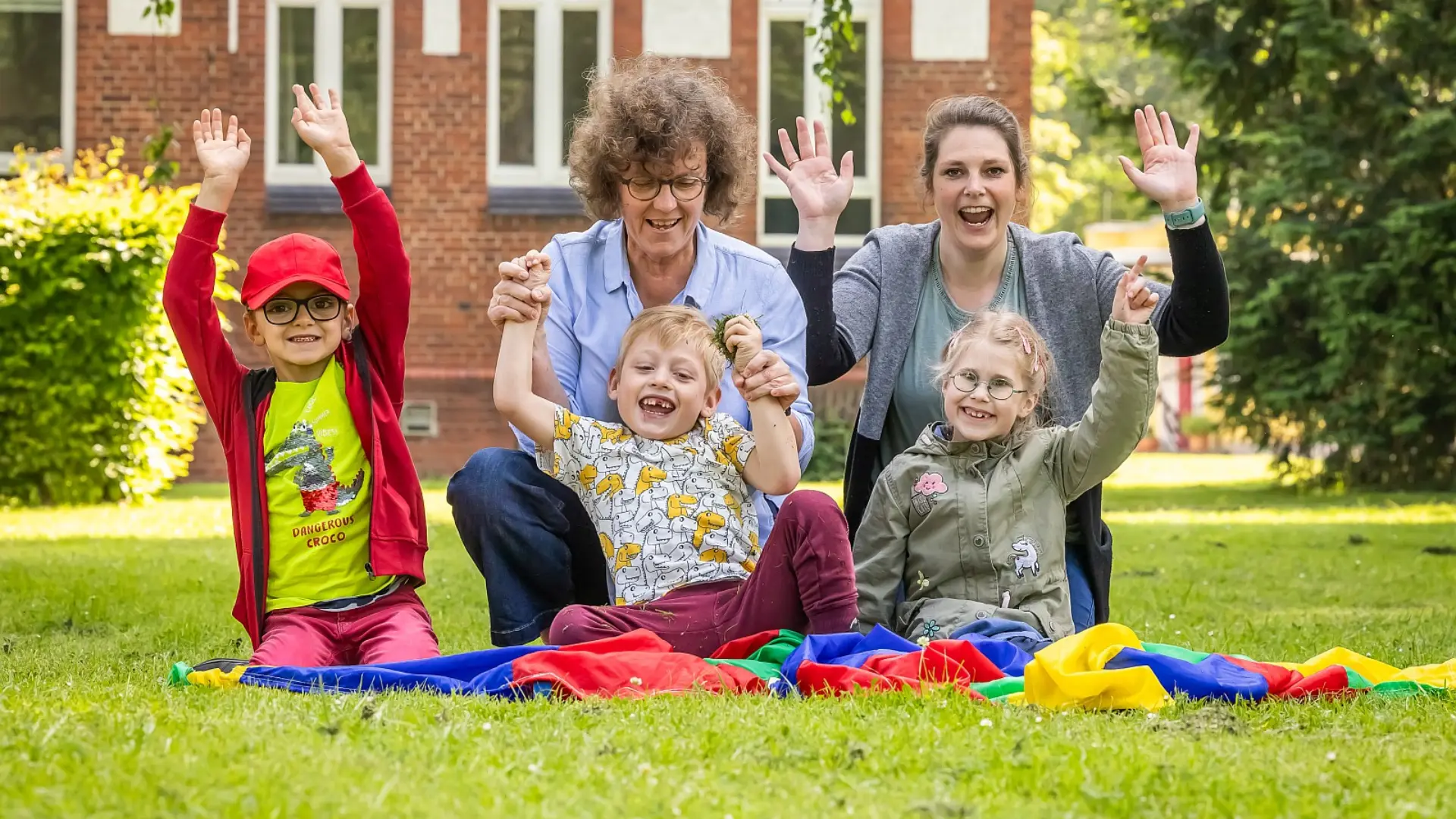 Zwei Pädagoginnen sitzen auf der Wiese mit drei Kindern im Vorschulalter, sie gucken in die Kamera und jubeln fröhlich mit hochausgestreckten Armen