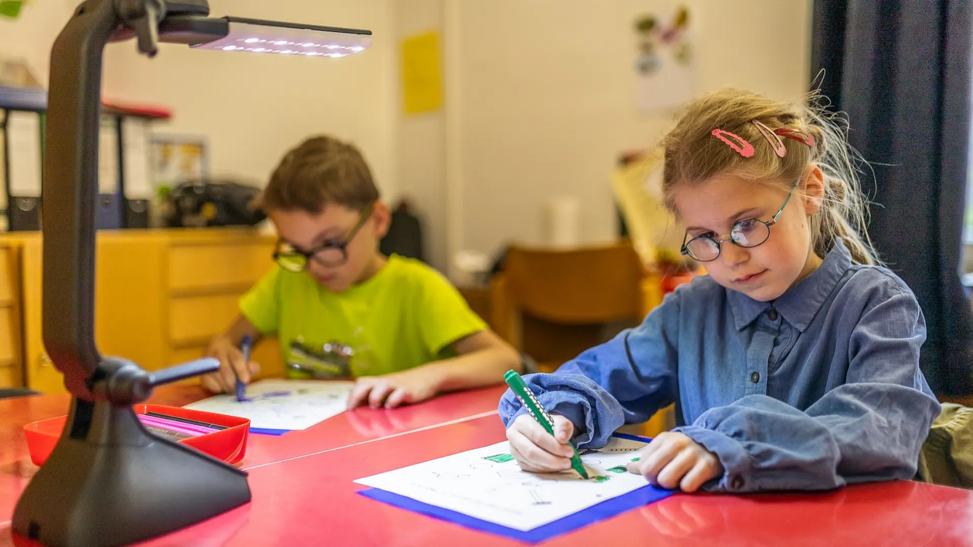 Zwei Kinder im Vorschulalter sitzen am Schreibtisch, leicht voneinander entfernt und füllen etwas in ihren Aufgabenheften aus. Ihre Arbeitsplätze sind mit Schreibtischlampen ausgeleuchtet und die Kinder sind in ihren Aufgaben vertieft.