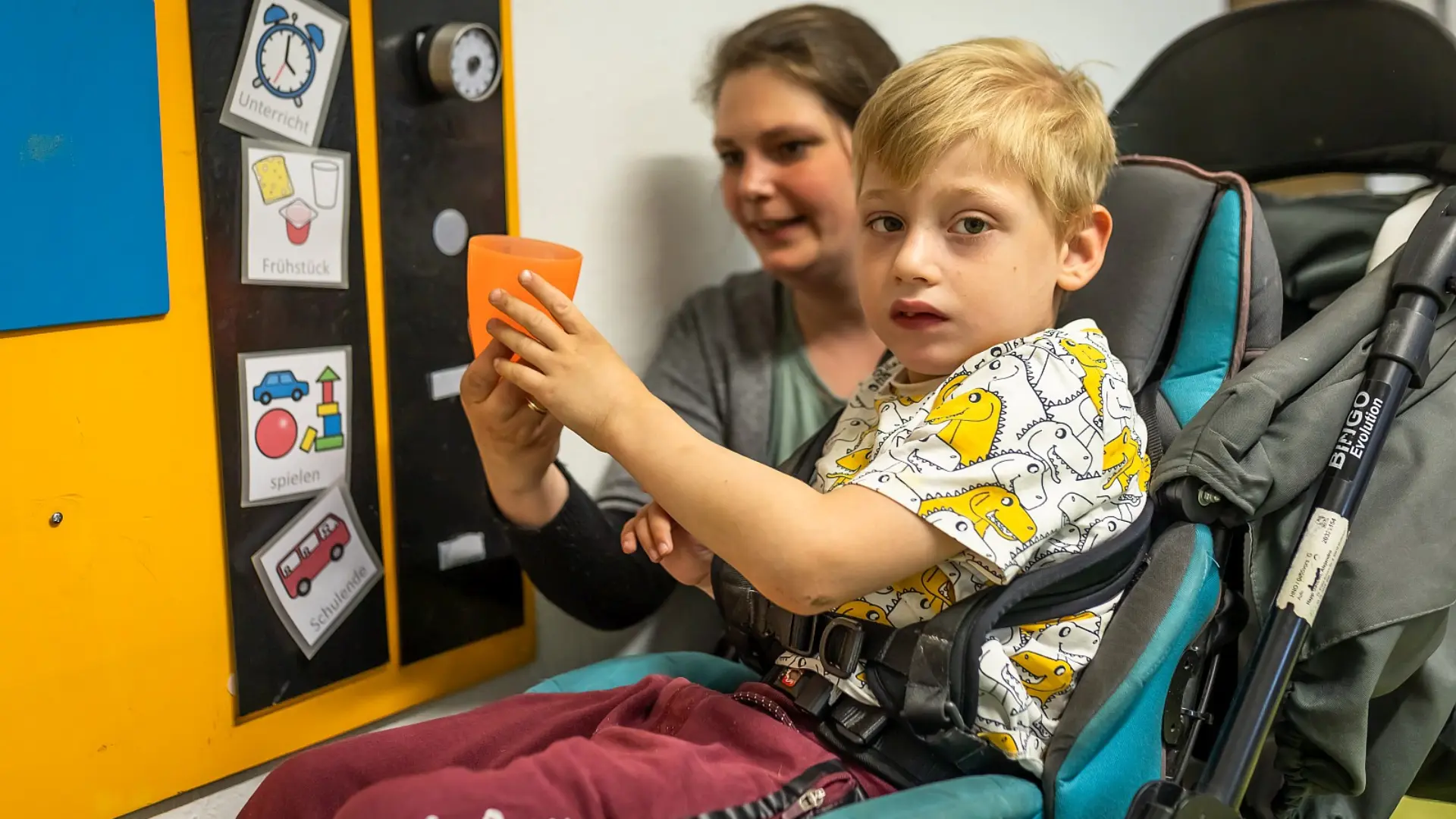 Ein Junge im Vorschulalter sitzt in einem gepolsterten Kinderwagen im Flur der Förderschule. Eine Lehrkraft reicht ihm einen Getränkebecher, er schaut zur Kamera. Beide Personen befinden sich vor einer Tafel mit Metacom-Symbolen.