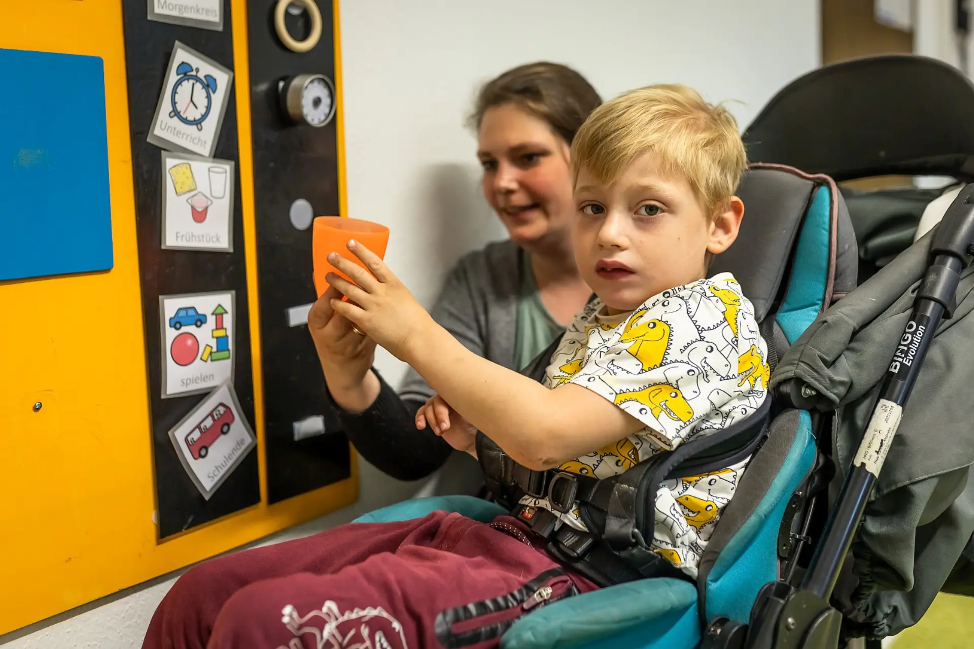 Ein Junge im Vorschulalter sitzt in einem gepolsterten Kinderwagen im Flur der Förderschule. Eine Lehrkraft reicht ihm einen Getränkebecher, er schaut zur Kamera. Beide Personen befinden sich vor einer Tafel mit Metacom-Symbolen.